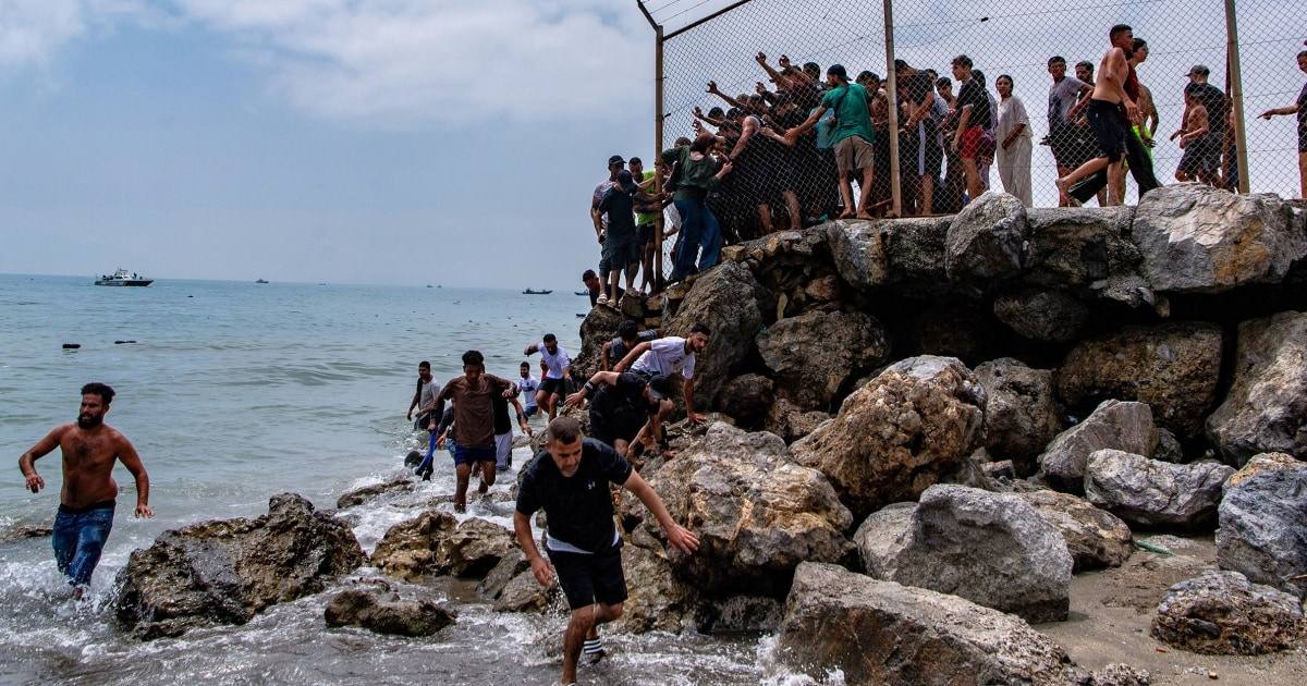 People at the rocky shoreline near the Ceuta border crossing during the July 2026 mass crossing from Morocco, with the border fence above them