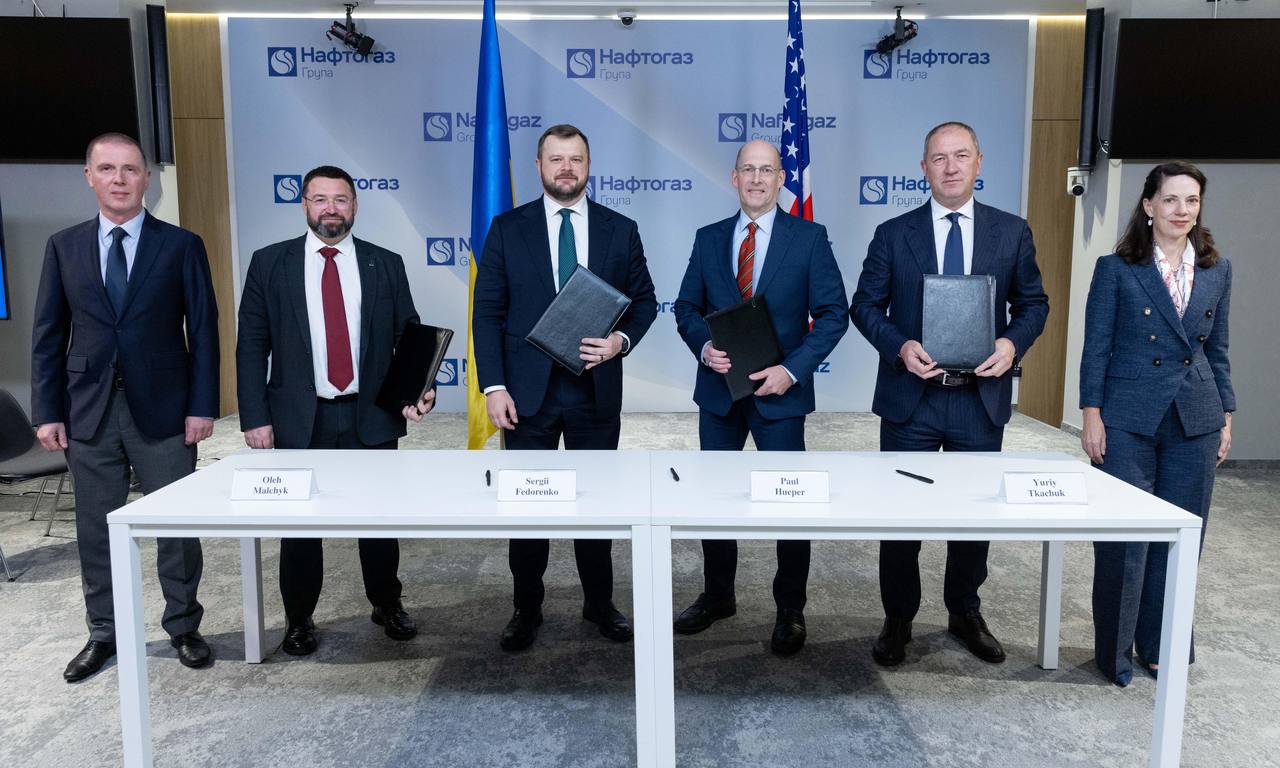 Six officials standing behind a signing table with Ukrainian and US flags and a Naftogaz backdrop after the memorandum signing on October 28, 2026; nameplates for Sergii Fedorenko and Paul Hueper are visible