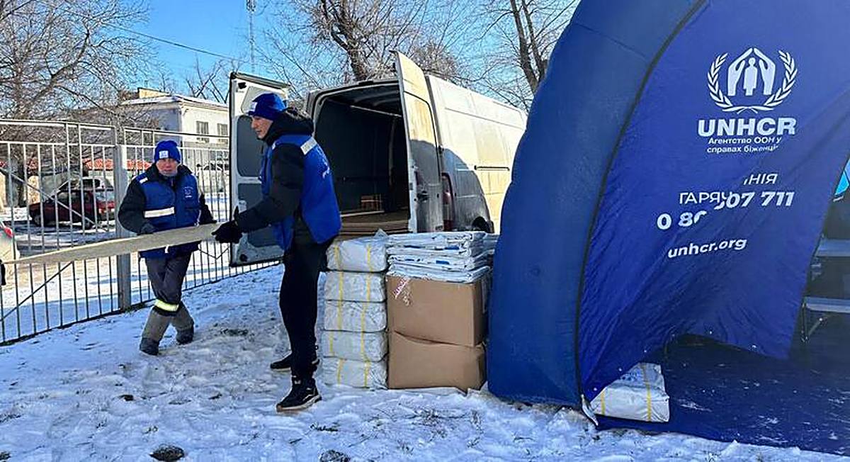 UNHCR aid workers unloading winter relief supplies beside a blue UNHCR tent in snow in Ukraine