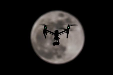 A quadcopter drone silhouetted against the full moon at night