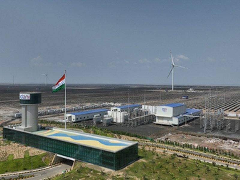 An Adani renewable energy plant in India with a large Indian flag, wind turbine and solar fields under a clear sky