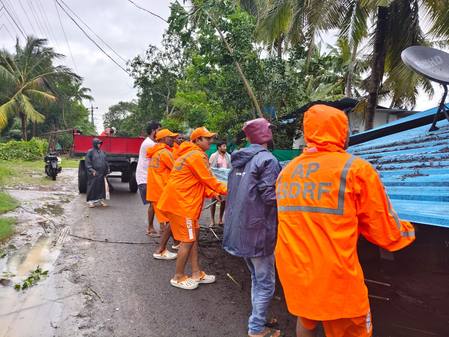 AP SDRF relief personnel in orange rain gear clearing a fallen tree from a road in cyclone-hit Andhra Pradesh