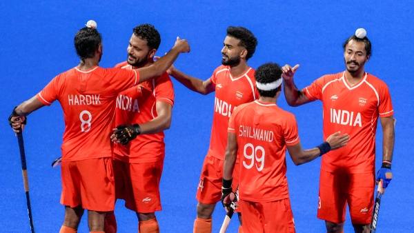 Indian men's hockey players in red kits celebrating a goal on a blue turf pitch