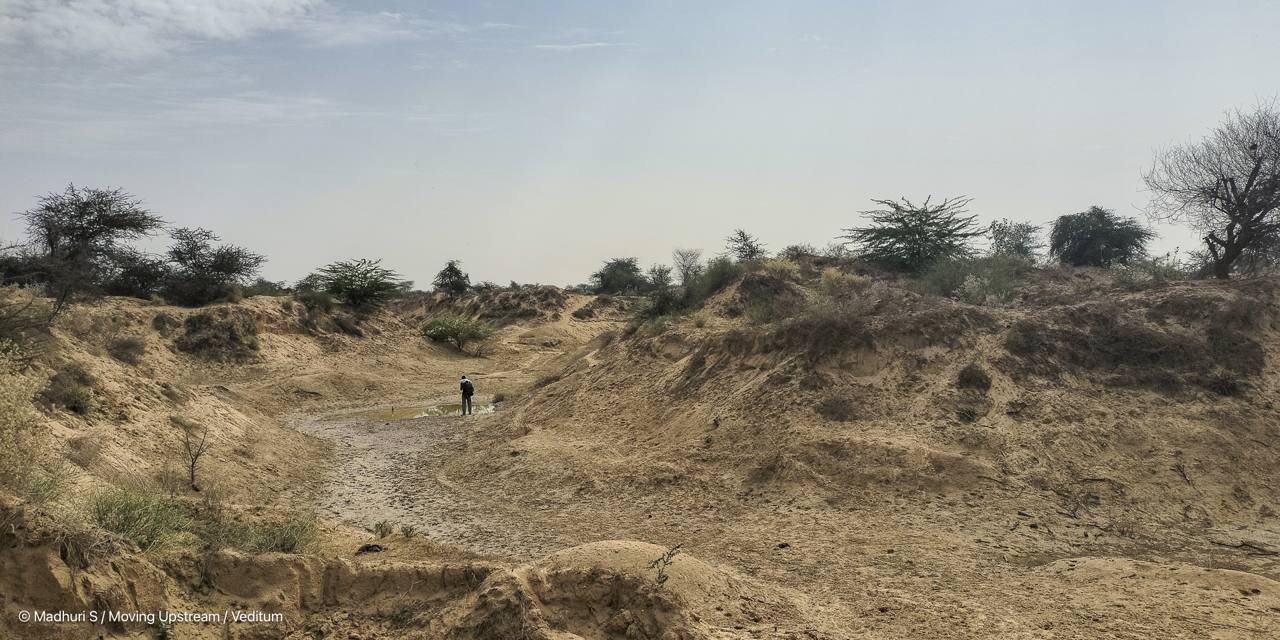 A dry, sandy stretch of the Luni riverbed in Rajasthan, with a lone figure standing near a small pool of water