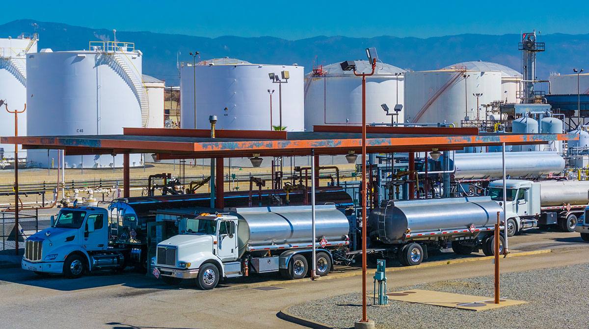 Fuel tanker trucks loading at a diesel storage terminal with large white storage tanks behind them under a blue sky