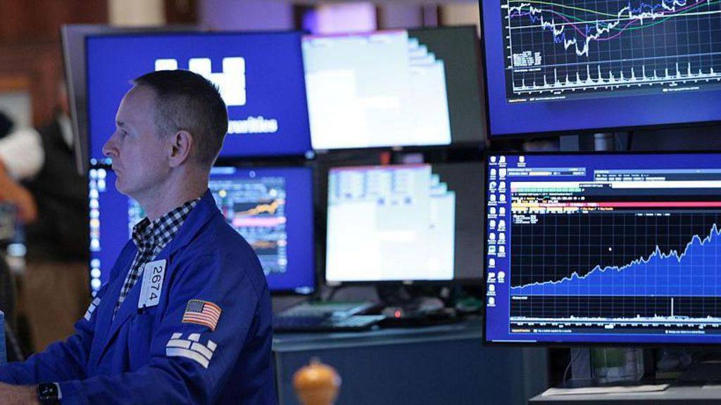 A trader in a blue jacket watching market screens with rising charts on the New York Stock Exchange floor